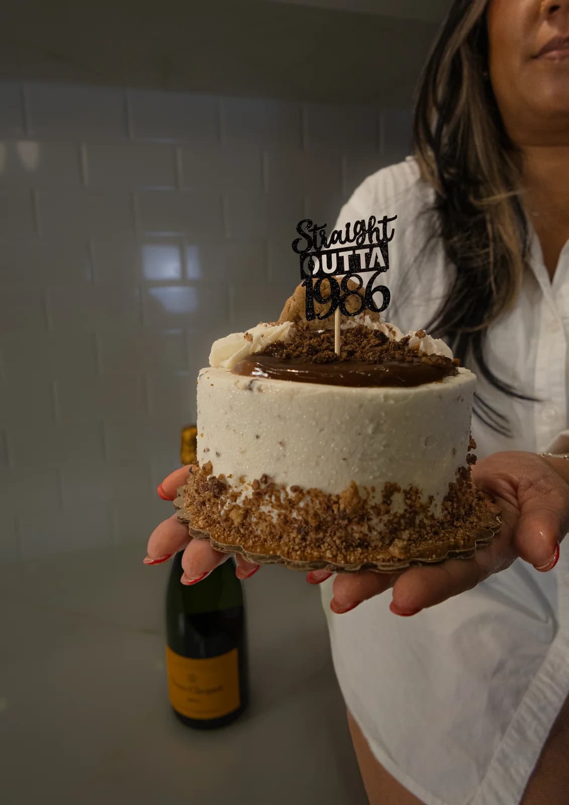 Close-up of hands with red nails holding a caramel-topped birthday cake with a "Straight Outta 1986" topper, a champagne bottle in the background.