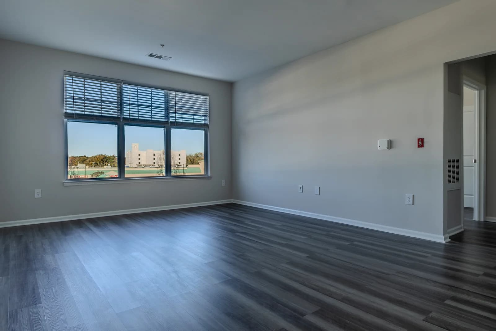 Empty living room with gray plank floors, white walls, and a wide window with blinds letting in natural light.
